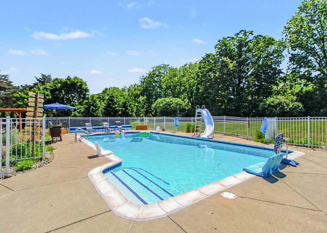 The inground pool with a built-in slide, surrounded by white fencing and a stone-paved deck under a clear blue sky.
