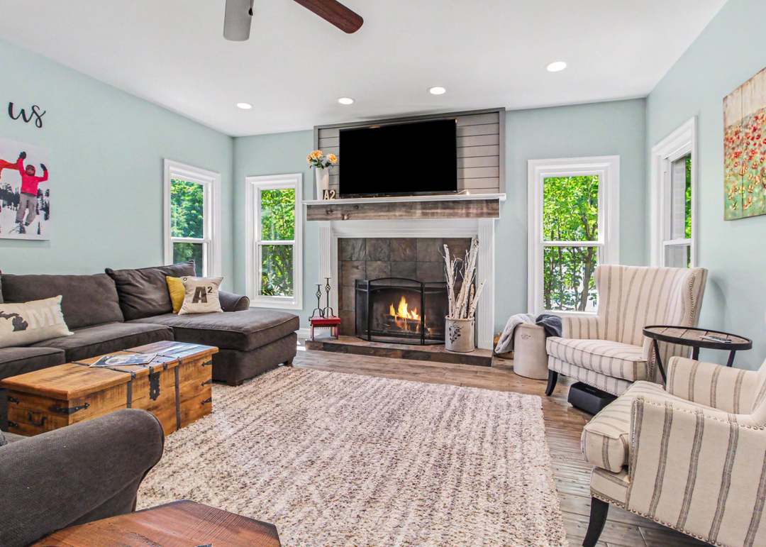 The living room with a lit gas fireplace under a shiplap-and-wood mantel, framed by striped wing chairs and tall windows.