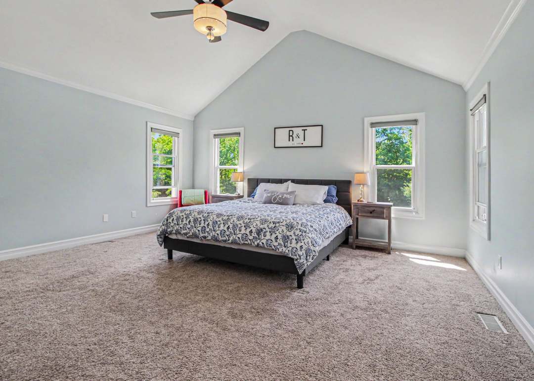 The primary bedroom with a vaulted ceiling, pale blue walls, three windows looking into the trees and soft carpet.