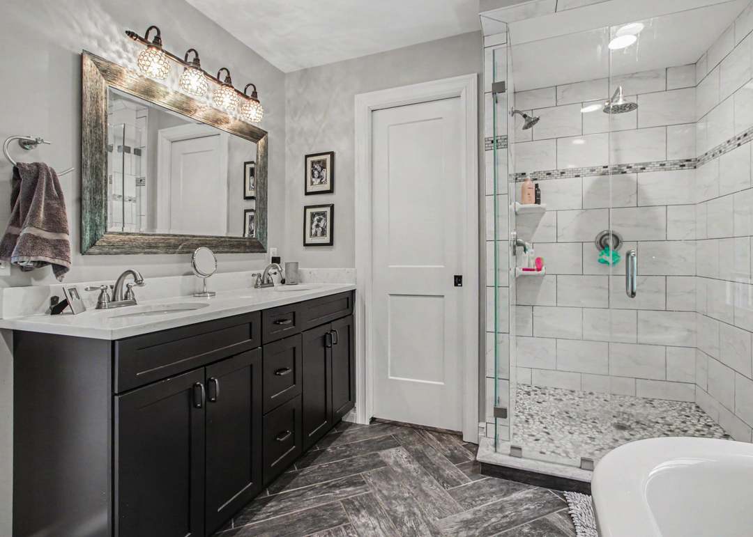 The primary bathroom with a double espresso vanity, a glass-walled tiled shower, a freestanding soaking tub and herringbone tile flooring.