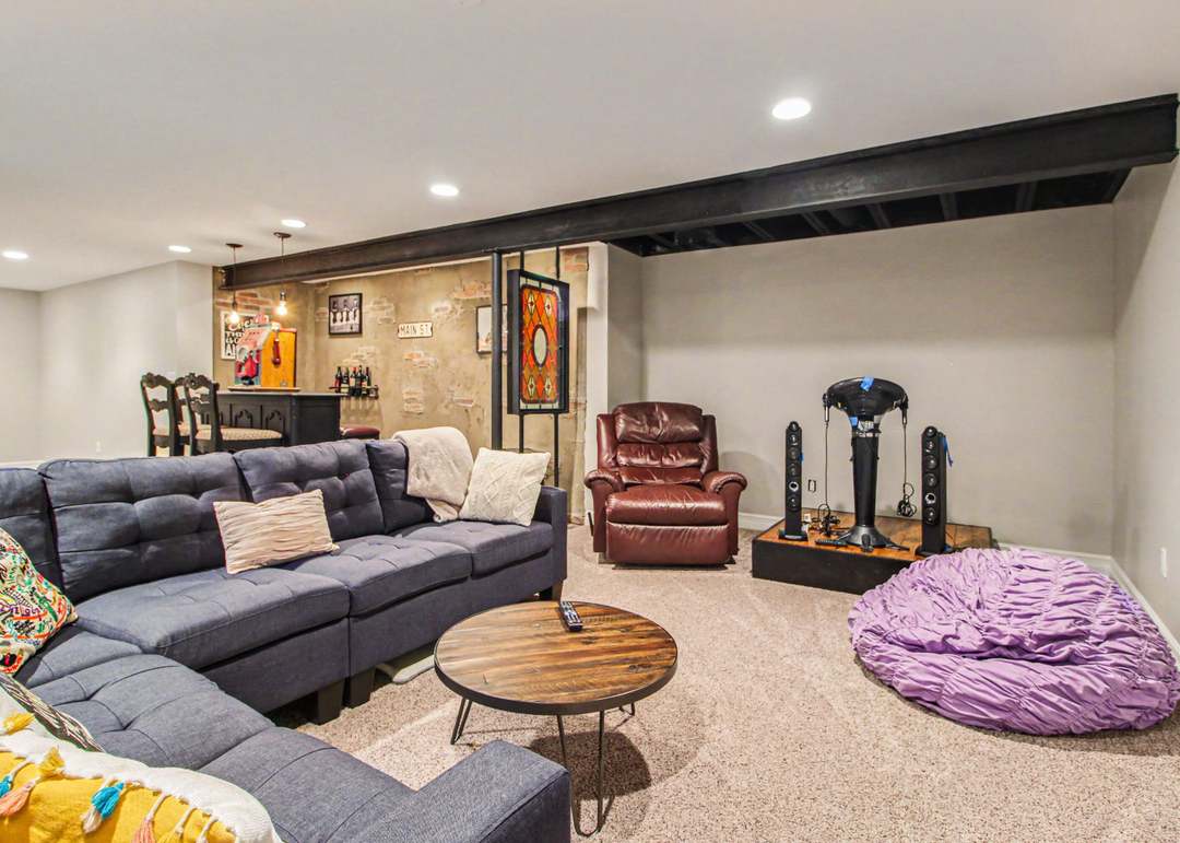 The finished lower level with a gray sectional, an exposed painted ceiling beam and a brick bar nook at the far end.