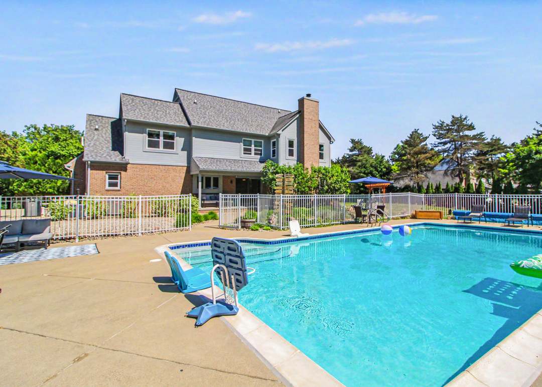 The rear of the house, brick and gray siding with a chimney, seen across the bright turquoise water of the fenced inground pool and its paver deck.